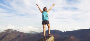 Young happy woman hiker with backpack standing on mountain peak with open arms